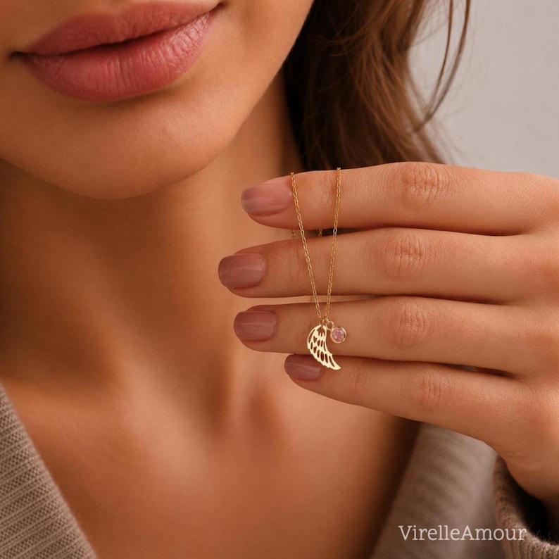 May include: A delicate gold necklace featuring a wing pendant with intricate detailing and a small, round pink gemstone. The necklace is displayed against a soft, neutral background, highlighting its elegant design. The brand name "VirelleAmour" is visible at the bottom right.