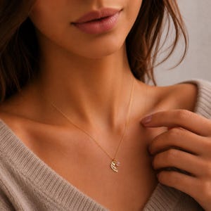 May include: A delicate gold necklace featuring a wing pendant and a small pink gemstone charm. The necklace is worn on a person and is a fine piece of jewelry. The gold chain is thin and the pendant is detailed.