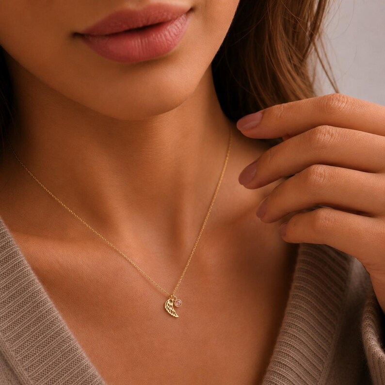 May include: A delicate gold necklace featuring a small wing pendant and a round, clear gemstone. The necklace is worn on a person's neck, with the chain and pendants in sharp focus. The background is blurred, highlighting the jewelry.