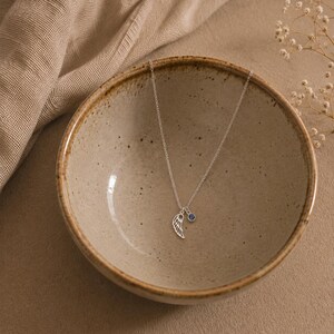 May include: A delicate silver necklace featuring a wing pendant and a small blue gemstone charm. The necklace is displayed inside a ceramic bowl with a neutral color palette, creating a minimalist aesthetic. The wing pendant is intricately detailed.