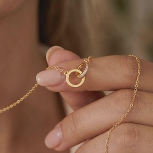 Puede incluir: Un delicado collar de oro con dos círculos entrelazados, uno adornado con pequeñas piedras brillantes. El collar tiene una fina cadena. La joya es sostenida por una persona con una manicura de tonos suaves y neutros. El collar está diseñado para llevarse al cuello.