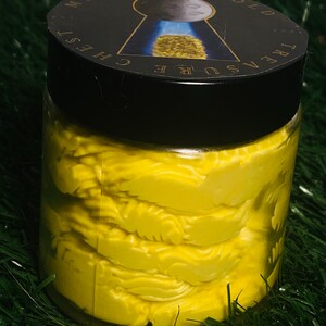 Op de afbeelding: Een glazen pot gevuld met een heldergele, getextureerde, romige substantie. De pot heeft een zwarte deksel met een gouden sticker waarop "Treasure Chest Midnight Gold" staat.