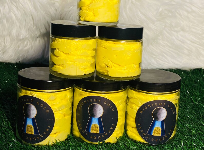 Op de afbeelding: Vijf glazen potten gevuld met gele body butter, gestapeld in een piramidevorm. Elke pot heeft een zwart deksel en een etiket met de tekst "Midnight Gold Treasure Chest".