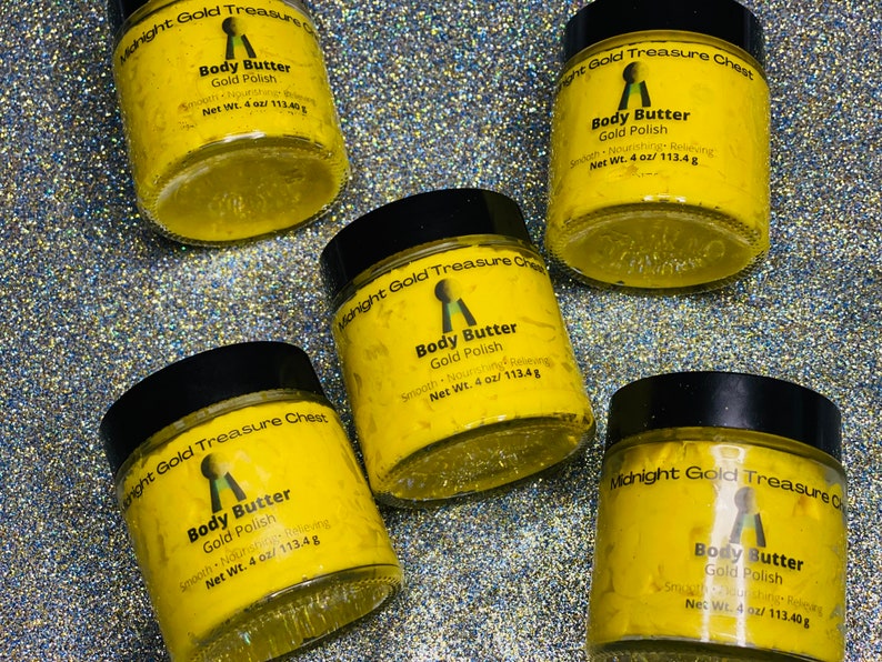 Op de afbeelding: Vijf gele potten met body butter en zwarte deksels. Elke pot heeft een gouden sleutel logo en de tekst "Midnight Gold Treasure Chest" en "Body Butter Gold Polish" met een nettogewicht van 4 oz/113,4 g.