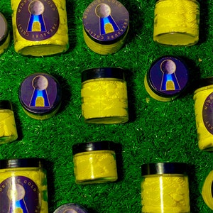 Op de afbeelding: Meerdere glazen potten met gele cr&egrave;me erin. De potten hebben zwarte deksels en een goud-blauw etiket met de tekst "Midnight Gold Treasure Chest". De potten staan op een groene ondergrond.