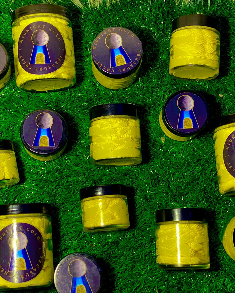 Op de afbeelding: Meerdere glazen potten met gele cr&egrave;me erin. De potten hebben zwarte deksels en een goud-blauw etiket met de tekst "Midnight Gold Treasure Chest". De potten staan op een groene ondergrond.