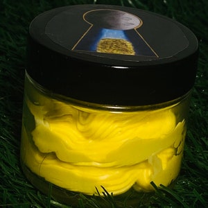 Op de afbeelding: Een gele pot met een zwart deksel. Het deksel heeft een sticker met een sleutelgat en een maan. De pot is gevuld met een gele substantie.