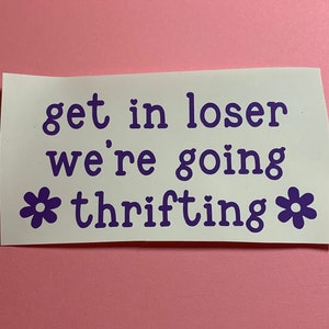 May include: A white sticker with purple text that reads "get in loser we're going thrifting." The text is in a playful font, with a heart before "loser" and flower graphics on either side of "thrifting."