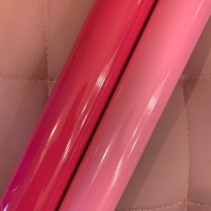 May include: Two rolls of glossy vinyl in shades of pink and red-pink. The rolls are side-by-side, with the darker roll on the left and the lighter roll on the right. The vinyl has a shiny, reflective surface, and the background is a quilted, light pink material.