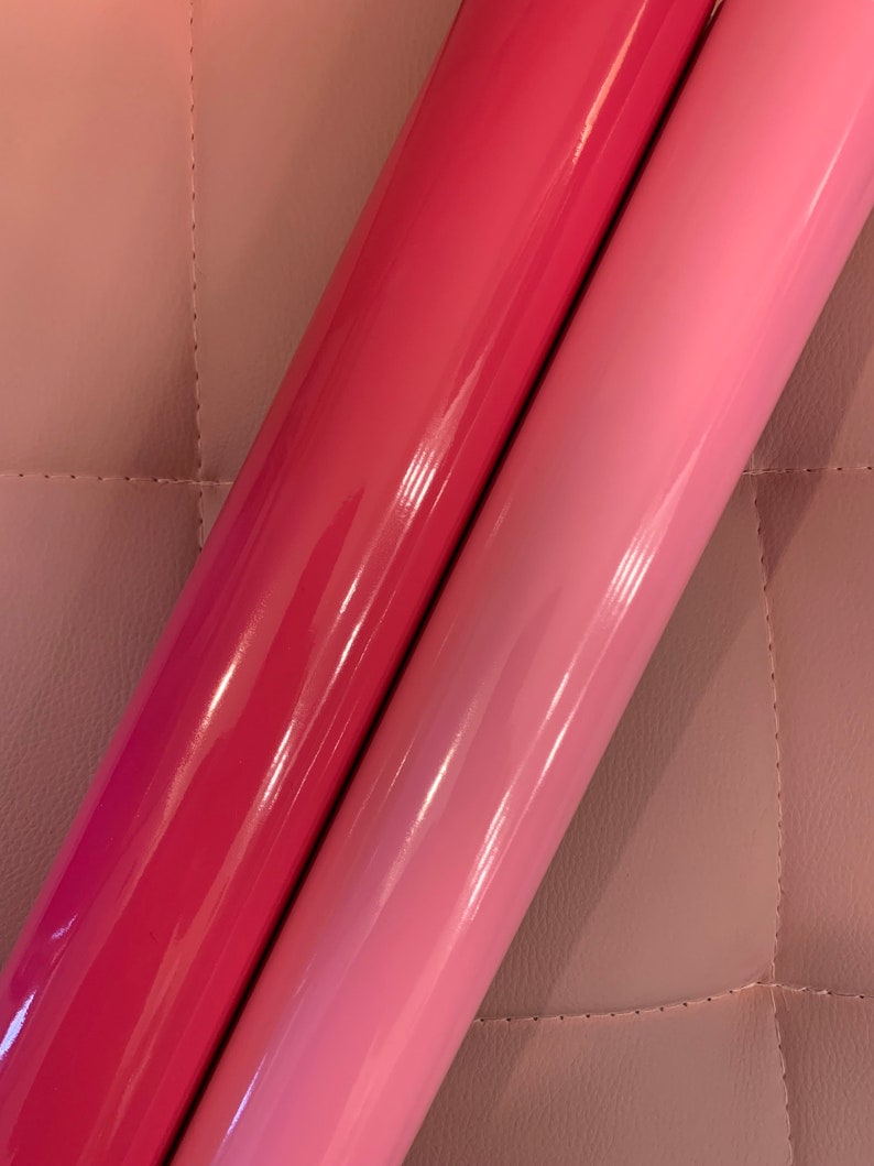 May include: Two rolls of glossy vinyl in shades of pink and red-pink. The rolls are side-by-side, with the darker roll on the left and the lighter roll on the right. The vinyl has a shiny, reflective surface, and the background is a quilted, light pink material.
