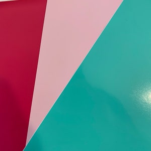 May include: An overhead shot of three sheets of colored paper. The colors are a deep red, a light pink, and a teal. The paper has a glossy finish and is arranged in a diagonal pattern.