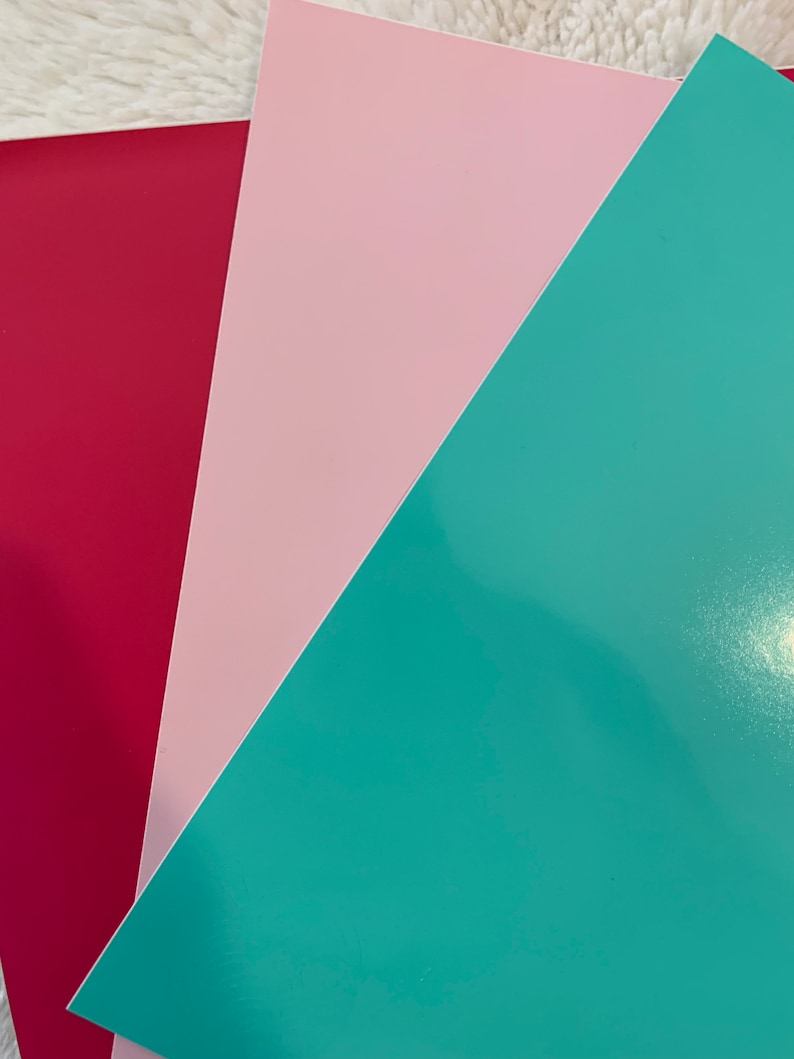 May include: An overhead shot of three sheets of colored paper. The colors are a deep red, a light pink, and a teal. The paper has a glossy finish and is arranged in a diagonal pattern.