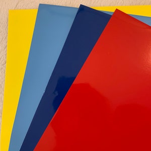 May include: A collection of glossy, colored sheets arranged in a fan shape. The colors include yellow, light blue, dark blue, and red. The sheets have a reflective surface, suggesting a vinyl or similar material.