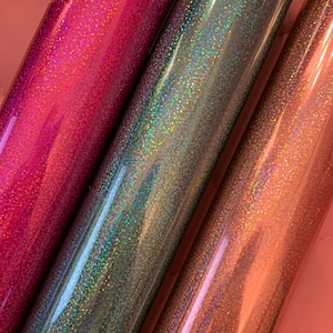 May include: Close-up of three rolls of glitter vinyl in shades of pink, teal, and rose gold. The vinyl has a holographic effect, reflecting multiple colors. The rolls are tightly wound and appear smooth and shiny, suitable for crafting projects.