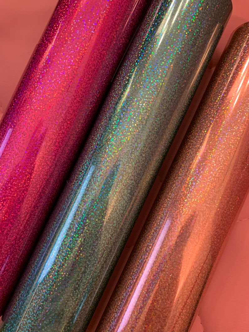 May include: Close-up of three rolls of glitter vinyl in shades of pink, teal, and rose gold. The vinyl has a holographic effect, reflecting multiple colors. The rolls are tightly wound and appear smooth and shiny, suitable for crafting projects.