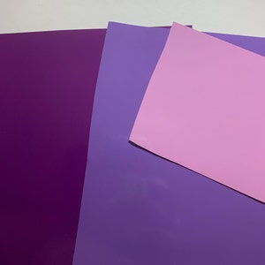 May include: An assortment of purple and pink sheets of paper. The sheets vary in shades, with a deep purple, a lighter purple, and a soft pink. The paper appears to be smooth and flat, suitable for crafts or design projects.