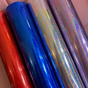 May include: Close-up of four rolls of glitter vinyl in red, blue, silver, and purple. The vinyl has a shiny, reflective surface with a speckled pattern. The rolls are tightly wound and appear to be new.