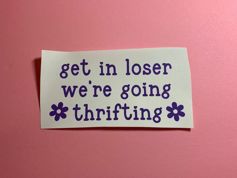 May include: A white sticker with purple text that reads "get in loser we're going thrifting." The text is in a playful font, with a heart before "loser" and flower graphics on either side of "thrifting."