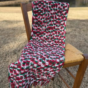 Può includere: Una coperta all'uncinetto fatta a mano in rosso, bianco e verde, drappeggiata su una sedia di legno. La coperta presenta un motivo strutturato e frange sul bordo. La sedia ha una seduta intrecciata e una struttura in legno scuro. La scena è ambientata all'aperto con uno sfondo sfocato.