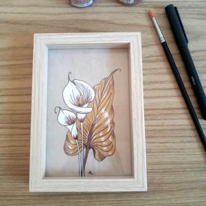 May include: A framed botanical art print featuring white calla lilies and a large, tan leaf. The artwork is in a light wood frame. Two bottles of metallic paint, a paintbrush, and a black pen are also visible.