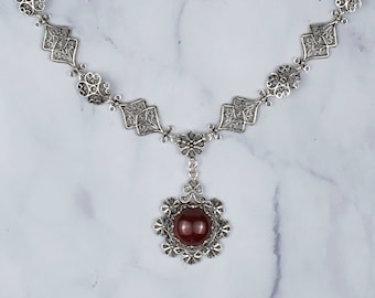May include: A silver filigree necklace with a red gemstone pendant. The necklace has a delicate chain and a small, intricate design.