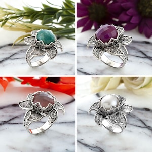 May include: Four silver rings with different gemstone centerpieces. The gemstones are green, purple, brown, and white. Each ring has a floral design with intricate details and small accent stones.