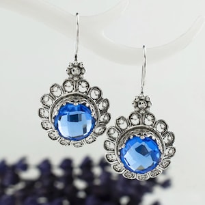 Op de afbeelding: Een paar zilverkleurige oorhangers met grote, facetgeslepen, blauwe edelstenen. De oorbellen hebben een bloemmotief rond de blauwe stenen en een decoratieve bovenkant. Ze hangen tegen een witte achtergrond.