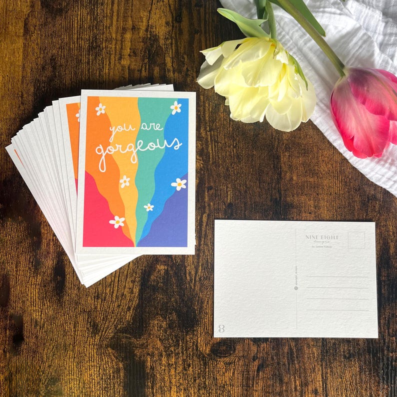May include: A stack of rectangular postcards featuring a colorful rainbow wave design with white daisy illustrations and the text you are gorgeous in white script. One single postcard is displayed face down showing a white back with address lines.