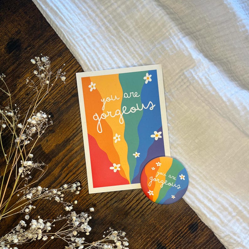 May include: A rectangular greeting card and a circular pinback button, both featuring a rainbow-colored wavy background with white daisy illustrations and the white text "you are gorgeous" printed in a cursive script on the front of each item.