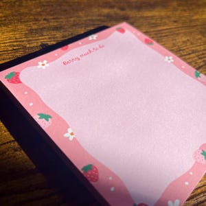 Könnte beinhalten: Ein quadratischer Notizblock mit einer hellrosa Mitte und einem gewellten rosa Rand, der mit einem Muster aus roten Erdbeeren, weißen Blumen und kleinen weißen Punkten verziert ist. Der Text Berry much to do steht in Rot oben links.