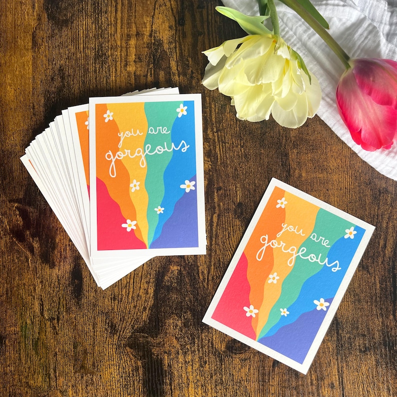 May include: A stack of rectangular greeting cards and one single card featuring a colorful vertical wavy rainbow pattern with white daisy flowers and the white handwritten text you are gorgeous printed on the front of each card.