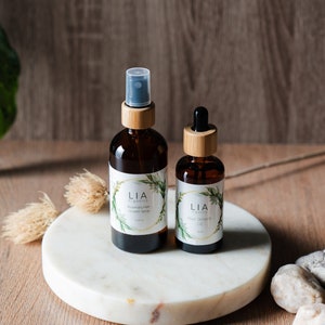 May include: Two amber glass bottles with bamboo lids and labels that read "LIA organics". One bottle is a spray bottle, the other has a dropper. The bottles sit on a white marble tray. The labels have a green leaf design.