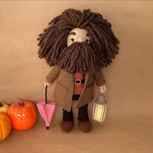 Può includere: Una bambola all'uncinetto con capelli e barba marroni, che indossa un cappotto marrone e tiene un ombrello rosa e una piccola lanterna. La bambola è in piedi davanti a tre zucche.