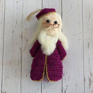 Può includere: Un giocattolo di peluche all'uncinetto di un mago che indossa una tunica viola con bordi dorati, una barba bianca e occhiali rotondi. Il giocattolo ha un fiocco dorato sul cappello.