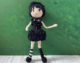 May include: A handmade crochet doll with black hair and a black dress. The doll's dress features a ruffled skirt and a high neckline. One leg has black and white striped stockings, while the other leg is solid black. The doll has a neutral skin tone.