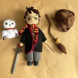 May include: A crocheted Harry Potter doll with a brown hat, a red and gold scarf, and a brown and gold broom. The doll is holding a wand and is standing next to a white crocheted owl.
