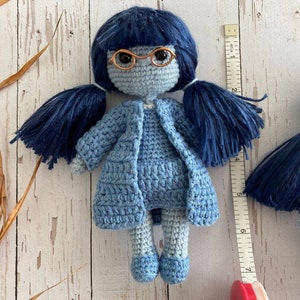 Può includere: Due bambole all'uncinetto con capelli in lana blu e vestiti blu. La bambola a sinistra indossa occhiali e un cappotto blu. La bambola a destra indossa un vestito blu. Entrambe le bambole sono alte circa 18 cm.