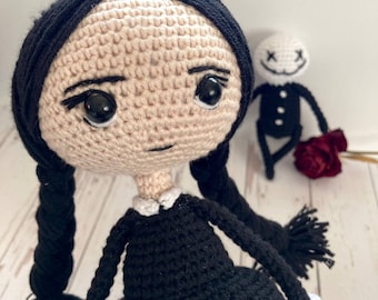 May include: A crocheted doll with black hair in pigtails, wearing a black dress with a white collar. The doll has a neutral expression with black eyes and a small, crocheted doll with a white face and black eyes standing behind it.