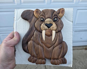May include: A wooden sculpture of a saber-toothed squirrel. The squirrel is brown with a dark brown tail and features two large white tusks. The sculpture is made of various wood pieces and is held on a piece of cardboard.