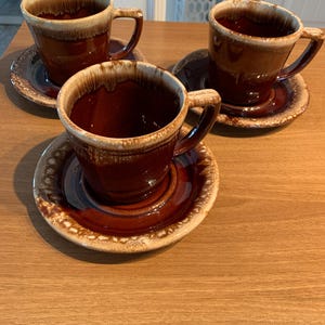 Puede incluir: Tres tazas de café de cerámica marrón con platillos a juego. Las tazas y los platillos tienen un acabado brillante con una combinación de colores de dos tonos, con un borde beige más claro y un cuerpo marrón más oscuro. Las tazas tienen asas.