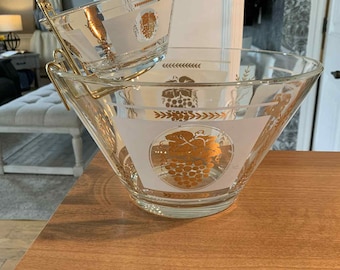 Puede incluir: Un juego de cristal transparente para patatas fritas y salsas con un soporte de metal dorado. El cuenco grande y el pequeño tienen una banda blanca con un diseño de uvas doradas. El juego está sobre una superficie de madera.
