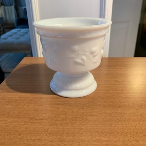 Puede incluir: Un cuenco blanco con pedestal y un diseño floral decorativo. El cuenco tiene una parte superior ancha y redondeada y una base con pie. Está hecho de un material brillante y opaco. El cuenco está sobre una superficie de madera.