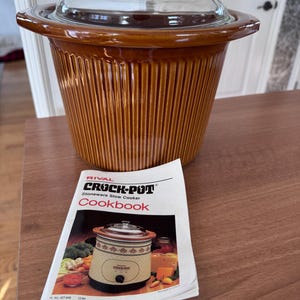 Może przedstawiać: Brązowy wolnowar Rival Crock-Pot z kamionki ze szklaną pokrywą i książką kucharską. Wolnowar ma żebrowany wzór i przezroczystą szklaną pokrywę z pokrętłem. Książka kucharska zawiera zdjęcie Crock-Pot i warzyw.
