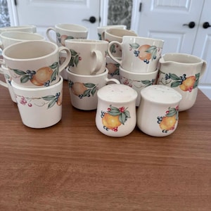 Könnte beinhalten: Eine Sammlung von cremefarbenen Keramikbechern, Tassen, einem Sahnekännchen sowie Salz- und Pfefferstreuern. Jedes Stück ist mit einem Fruchtdesign mit Orangen, Blaubeeren und grünen Blättern verziert. Das Set ist auf einer Holzoberfläche angeordnet.