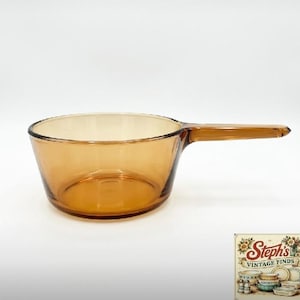 May include: A translucent amber-coloured glass saucepan featuring a round bowl and a single extended handle. The glass has a smooth, polished finish and a consistent warm brown hue throughout the entire structure of the cooking vessel.