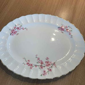May include: White oval serving platter with a scalloped edge and gold trim. The platter is decorated with pink cherry blossom branches. A decorative piece for serving food.