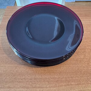May include: A stack of six dark blue glass plates with a red rim. The plates are round with a slightly raised edge and a smooth surface. The plates are stacked on a wooden surface.