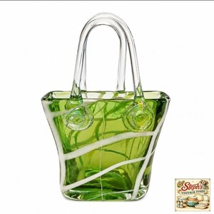 May include: A decorative glass handbag sculpture in a vibrant lime green hue. The piece features clear glass handles and a series of opaque white glass ribbons that wrap diagonally around the body of the bag, creating a distinct contrast.