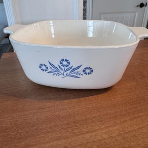 May include: A white, square casserole dish with a blue floral pattern. The dish has two handles and is made of ceramic. The blue floral design is centered on the front. The dish is on a wooden surface.