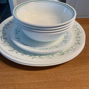 Puede incluir: Un conjunto de cuencos y platos de cerámica blanca con un diseño floral verde y azul. Los cuencos tienen un borde verde y azul. Los platos y cuencos están apilados sobre una superficie de madera.